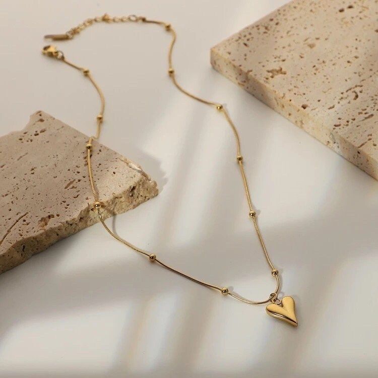 Gold heart pendant necklace on a stainless steel anti-tarnish chain displayed on a beige jewelry stand.