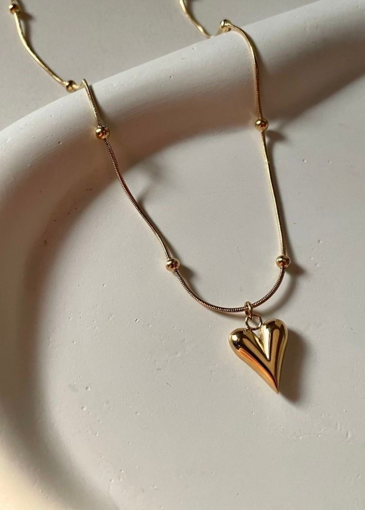 Gold heart pendant necklace on a stainless steel anti-tarnish chain displayed on a beige jewelry stand.