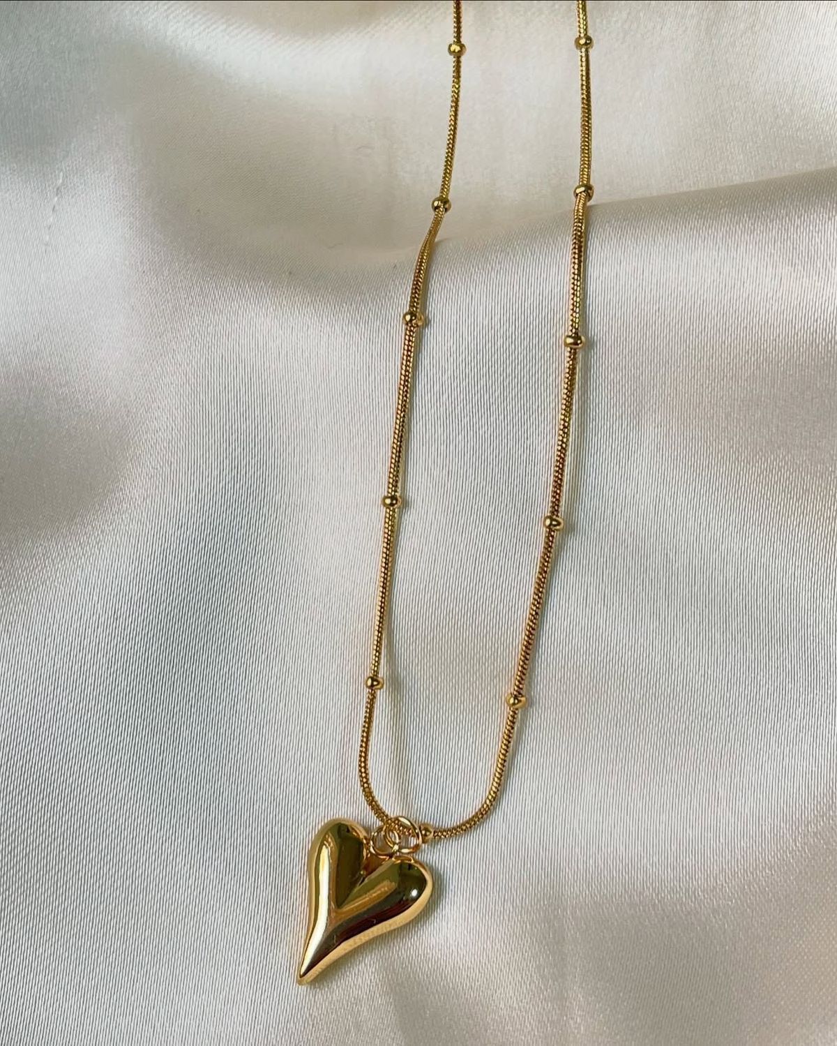 Gold heart pendant necklace on a stainless steel anti-tarnish chain displayed on a beige jewelry stand.